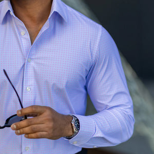 A man in COLLARS AND CO's Semi Spread Collar Apex Performance Dress Shirt in Lavender and Blue Multi Gingham adjusts his collar outdoors, surrounded by green plants and a blurred background.