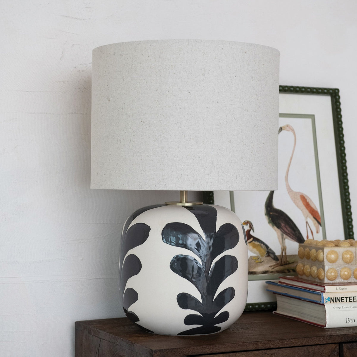 The CREATIVE COOP BLACK AND CREAM STONEWARE TABLE LAMP features a round ceramic base with black leaf designs and a light beige linen shade, set against a plain white background.