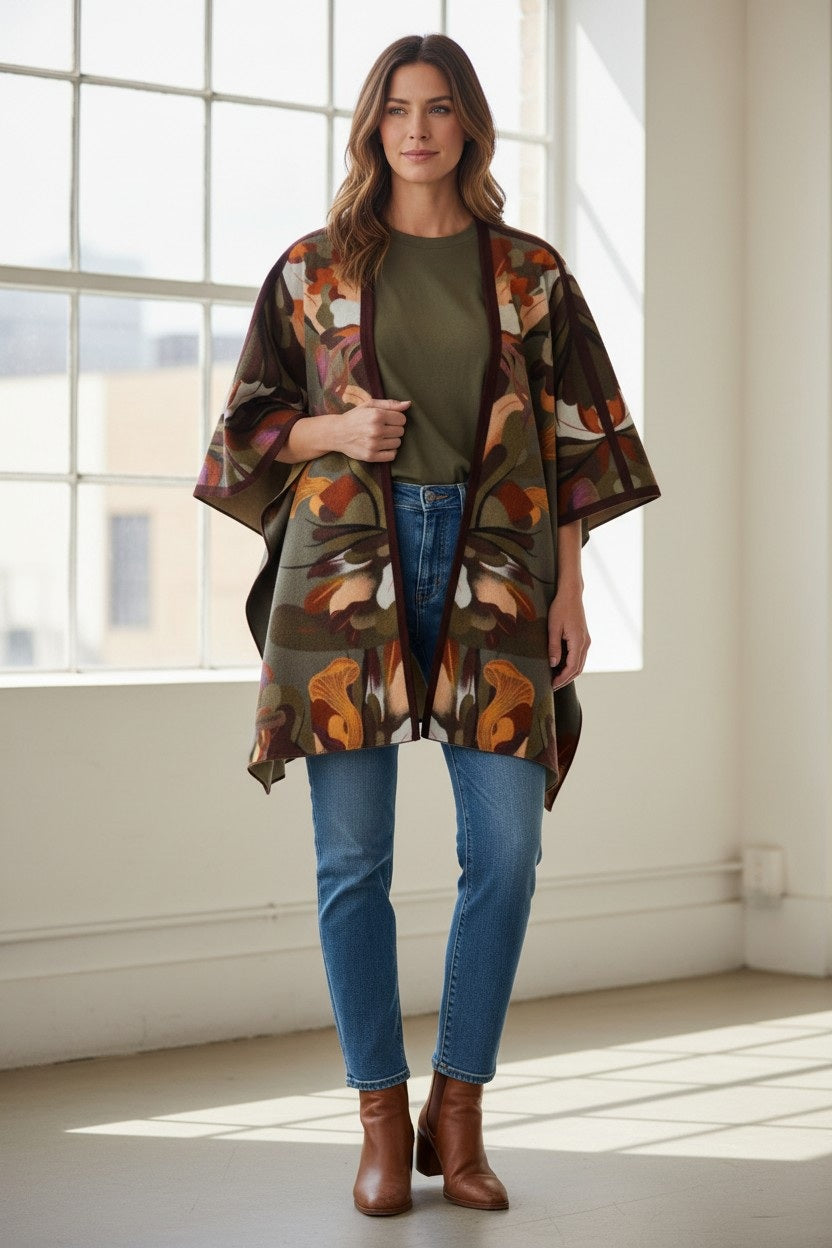 A woman stands indoors in front of large windows with natural light streaming in, wearing jeans, brown ankle boots, a green top, and the FRANCO FERRARI - WOOL CAPE by FRANCO FERRARI—a stylish piece perfect for lightweight layering.