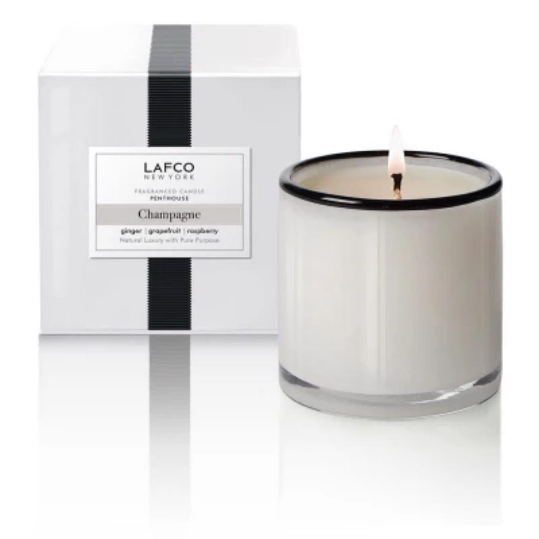 A lit white candle in a clear glass jar with a black rim is shown next to its sleek white packaging box. The box, adorned with a black vertical stripe, features the text &quot;LAFCO New York&quot; and &quot;Champagne, ginger, sparkling grapefruit, raspberry.&quot; This hand-poured LAFCO - CHAMPAGNE, PENTHOUSE 15OZ candle epitomizes the art of luxurious candles.