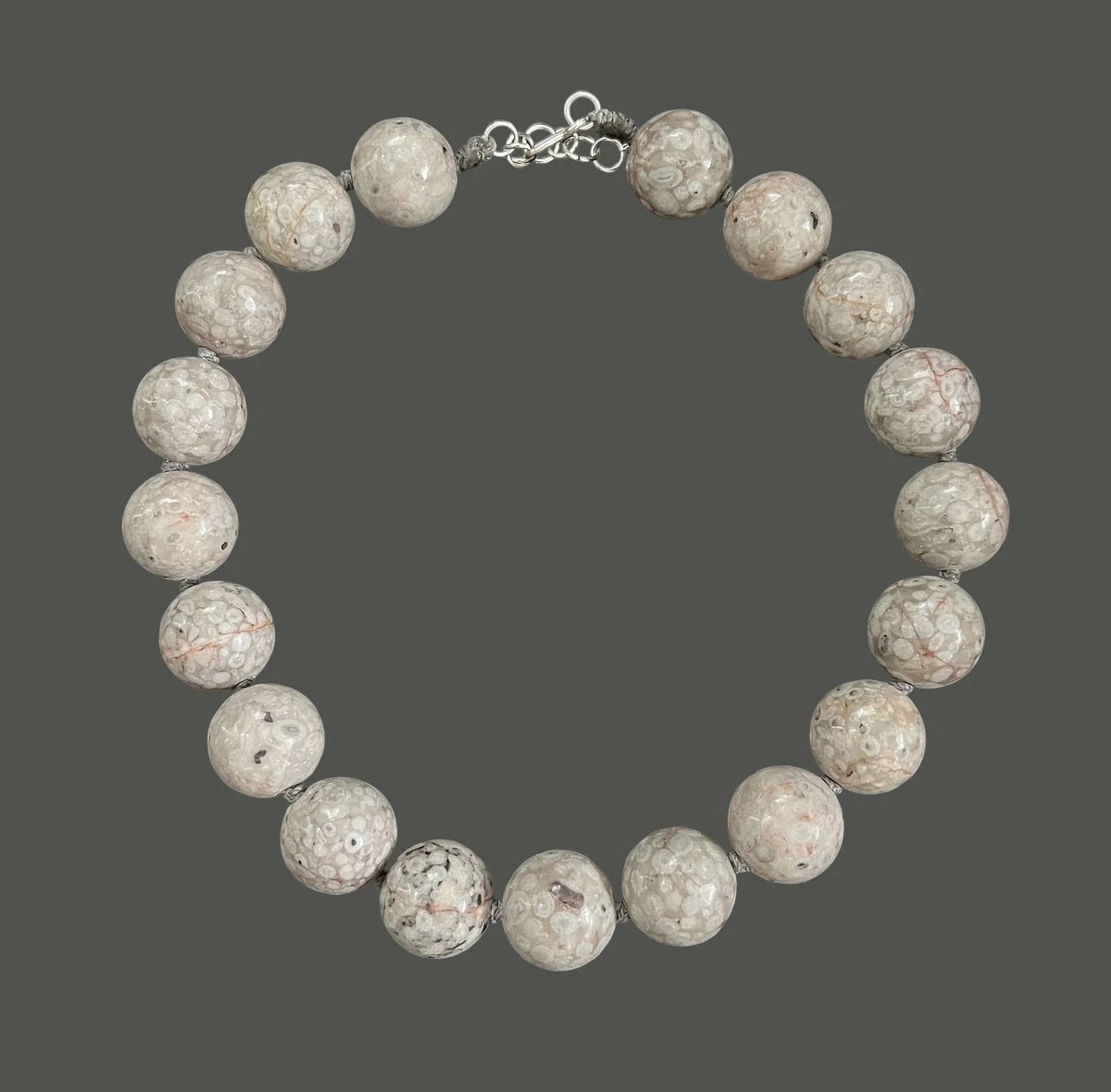 The ANN LIGHTFOOT Fossil Coral Ras du Cou Necklace features large, polished round fossil coral beads in light gray and beige with circular patterns and a sterling silver clasp, shown on a dark gray background.