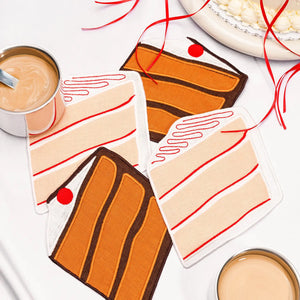 The LINEN CAKE COCKTAIL NAPKINS by PIECEWORK PUZZLES feature four embroidered cotton slices of classic layer cake, complete with cherry and icing details, resting alongside two coffee mugs and red ribbons.
