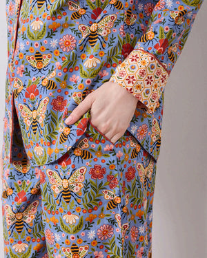 A woman with light brown hair sits against a white wall, wearing the PRINTFRESH - Bee Tapestry Long Pajama Set in Delphinium Blue. This organic cotton pajama set from PRINTFRESH features a colorful floral and bee pattern with vibrant red and yellow cuffs, collar, and pocket trim, beautifully blending playful design with cozy comfort.