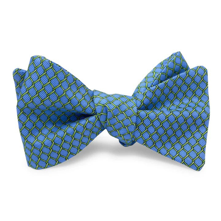 The BIRD DOG BAY PETER-BLAIR Fencing Silk Bow Tie in Blue showcases a classic butterfly shape with a blue base and small green and white geometric pattern, displayed flat on a white background.