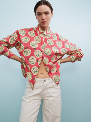 A woman sits on a clear chair with one arm propped on her head, wearing the 0039 ITALY LUCY BLOUSE IN A FRUIT PRINT, white pants, and yellow flats, against a light blue wall and gray carpet.