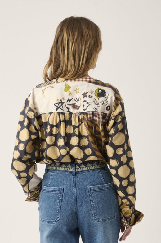 A person with curly hair stands indoors by a window, smiling softly with hands in pockets, dressed in blue jeans and the STORIATIPIC - MARTGOT BLOUSE IN DELTA by STORIATIPIC—a patterned cotton button-up featuring abstract designs. This blouse is easy to care for: simply machine wash gently at 30 degrees to keep it looking fresh.