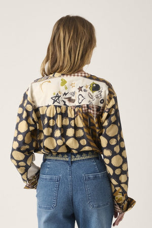 A person with curly hair stands indoors by a window, smiling softly with hands in pockets, dressed in blue jeans and the STORIATIPIC - MARTGOT BLOUSE IN DELTA by STORIATIPIC—a patterned cotton button-up featuring abstract designs. This blouse is easy to care for: simply machine wash gently at 30 degrees to keep it looking fresh.