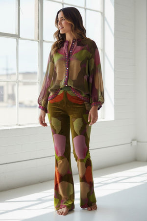A woman stands barefoot in a bright room, smiling, wearing the FRANCO FERRARI - KANADA VELVET FLARE TROUSER paired with a sheer, patterned blouse; both pieces feature earthy tones with pink, purple, and green abstract shapes.