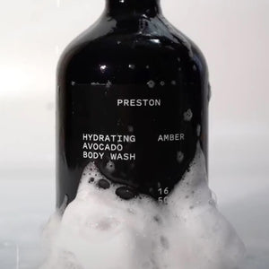 A black bottle of PRESTON - BODY WASH labeled "Hydrating Avocado Body Wash Leather" is centered against a beige background. Made in the USA with plant-based ingredients, this 16 oz (500 ml) moisturizing body wash from the brand PRESTON ensures hydrated skin without harsh chemicals.