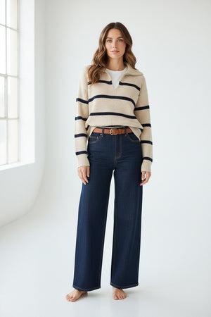A woman stands barefoot in a minimal, bright room, wearing the MERSEA Camden Sailor Sweater—a travel-ready knit with black horizontal stripes on a white base—paired with high-waisted wide-leg dark blue jeans and a brown belt.