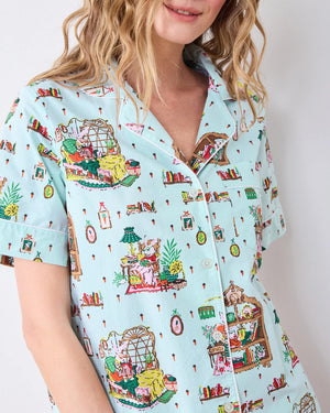 A person stands barefoot against a white wall, smiling and wearing the PRINTFRESH - READING NOOK PAJAMA SHORT SET by PRINTFRESH—colorful, organic cotton pajamas ideal for relaxing in your favorite reading nook.
