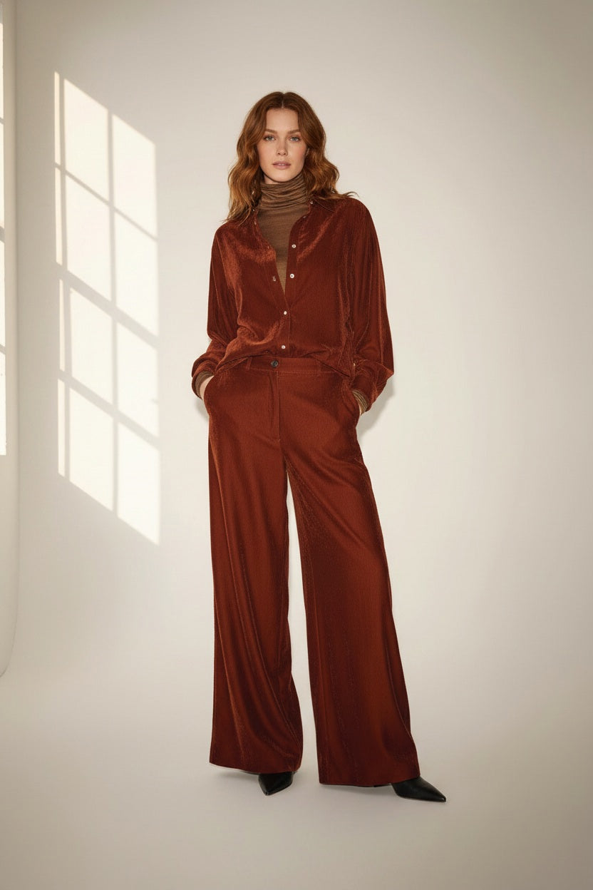 A person with long wavy hair stands indoors wearing a loose-fitting, rust-colored velvet jacket paired with the NUDE - WIDE LEG TROUSER IN TOBACCO by NUDE. Crafted from soft viscose baby cord, the high-rise trousers flatter their silhouette as they rest their hands in the jacket pockets. Soft natural light streams through a window, casting gentle shadows on the wall.