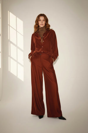 A person with long wavy hair stands indoors wearing a loose-fitting, rust-colored velvet jacket paired with the NUDE - WIDE LEG TROUSER IN TOBACCO by NUDE. Crafted from soft viscose baby cord, the high-rise trousers flatter their silhouette as they rest their hands in the jacket pockets. Soft natural light streams through a window, casting gentle shadows on the wall.