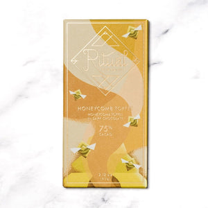 A gold and yellow RITUAL CHOCOLATE - HONEYCOMB TOFFEE CHOCOLATE BAR wrapper with geometric designs holds a 2.12 oz (60 g), 75% cacao single origin dark chocolate bar, displayed on a marble background.