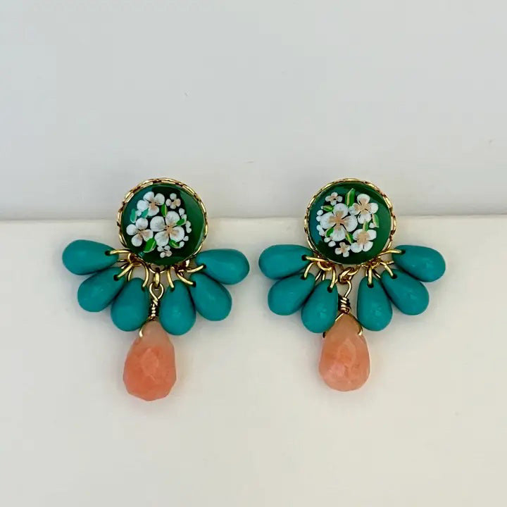The SANDRINE FRANCE STUDIO GRACIE EARRINGS feature colorful floral motifs with green, white, and gold tops, blue beads, and an orange teardrop bead. Handmade in NYC, they are displayed on a clear stand against a plain gray background.