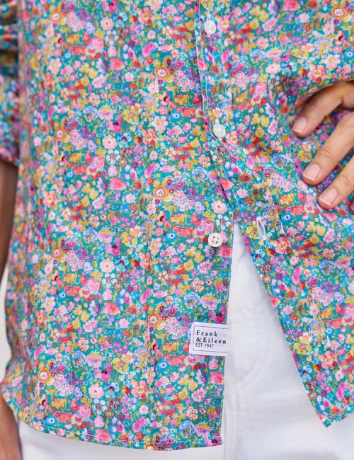 A woman with long brown hair smiles as she stands outside by a white wooden wall, wearing the FRANK & EILEEN EILEEN Relaxed Button Up in Italian Classic Linen Multicolor Flowers, paired with white pants and a straw tote bag nearby.