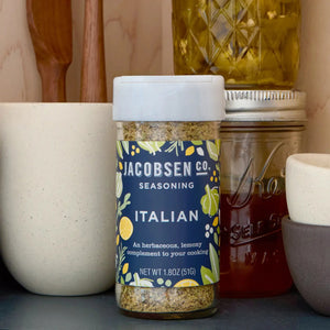 A jar of JACOBSEN SALT - ITALIAN SEASONING from JACOBSEN SALT CO. sits on a kitchen counter near utensils, a mason jar with a metal lid, and a ceramic cup. The spice jar label features a blue and yellow floral design.