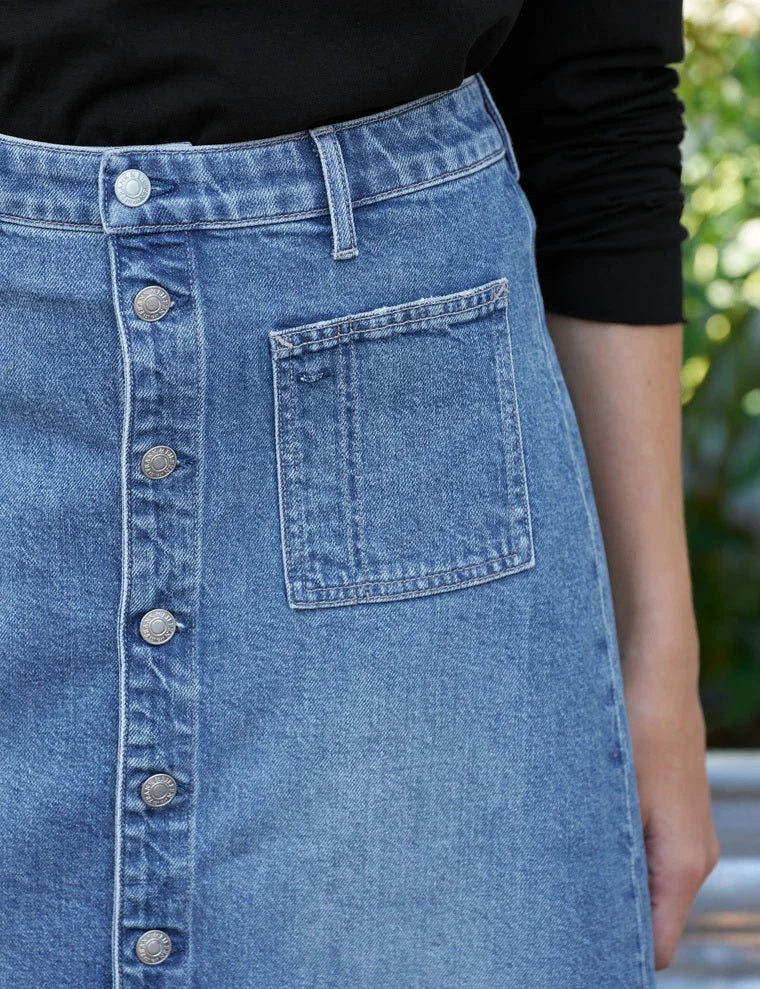 A woman walks on a dirt road near a fence, wearing a tan button-up shirt and the FRANK & EILEEN - SHANNON CLASSIC DENIM SKIRT IN 1978 WASH, paired with brown boots. In the background, trees and horses stand in a grassy field.