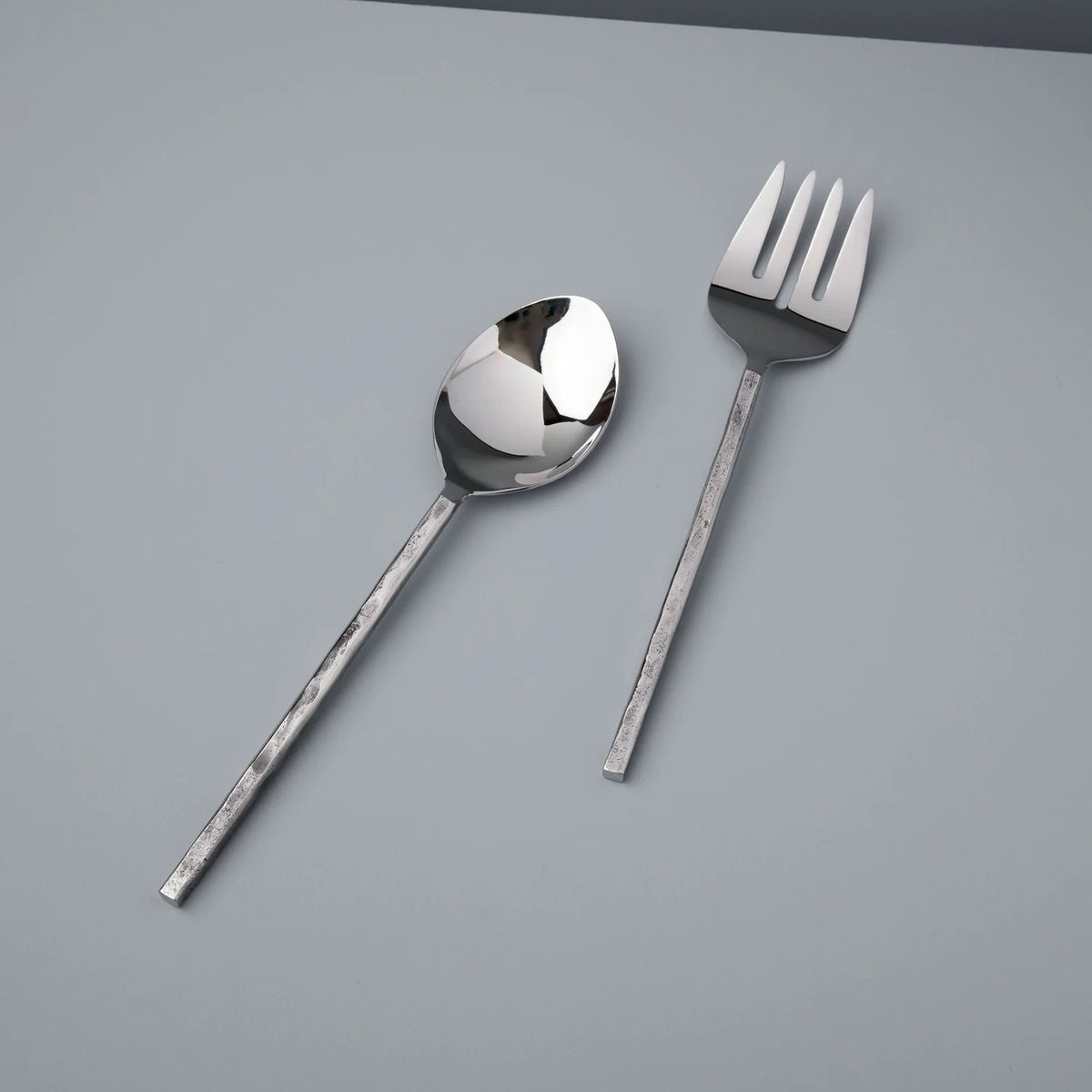 An AZURA SERVING SET by BE HOME, INC, consisting of a polished silver spoon and a three-pronged silver fork, is positioned on a light grey surface. Both utensils feature long, slender handles with an intricate textured finish and unique design. They are arranged parallel to each other but oriented in opposite directions. To maintain their delicate appearance, it is recommended to hand wash them with mild soap.