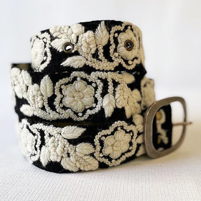 The JENNY KRAUSS Two Toned Belt in Black and Cream features intricate cream floral embroidery and a silver buckle, handmade in Peru. Shown coiled on a light background.