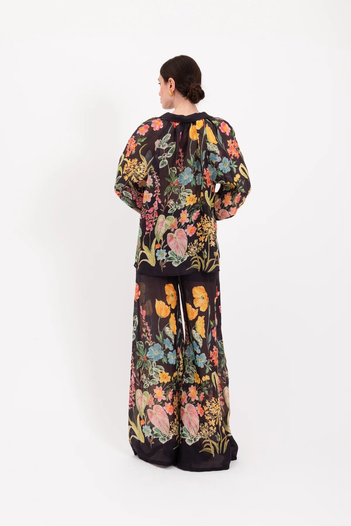 A person stands facing forward, wearing the LAVI - APHRODITE BOTANICAL BLOUSE by LAVI—a loose-fitting, long-sleeved statement blouse featuring a colorful floral pattern—against a plain white background.