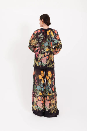 A person stands facing forward, wearing the LAVI - APHRODITE BOTANICAL BLOUSE by LAVI—a loose-fitting, long-sleeved statement blouse featuring a colorful floral pattern—against a plain white background.