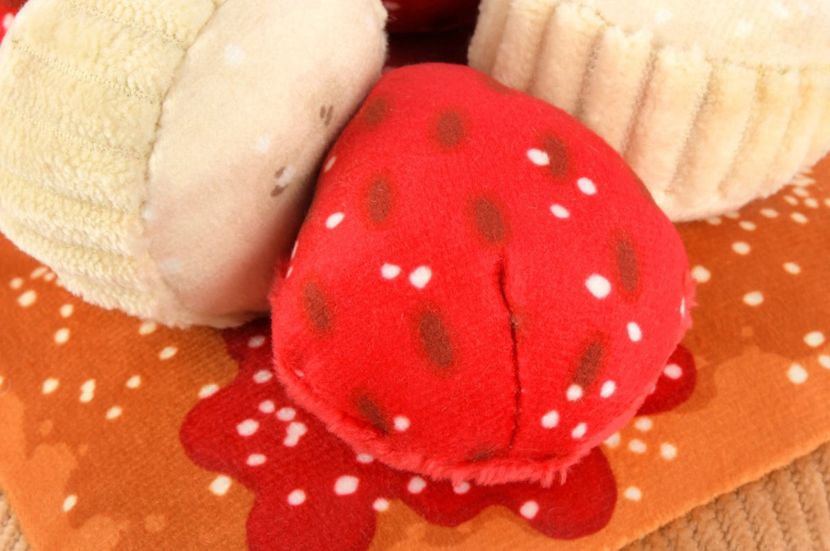 Close-up of the PLAY PET LIFESTYLE THICK ‘N FLUFFY FRENCH TOAST DOG TOY on a brown fabric with red and white patterns, pictured with plush strawberry and muffin toys.