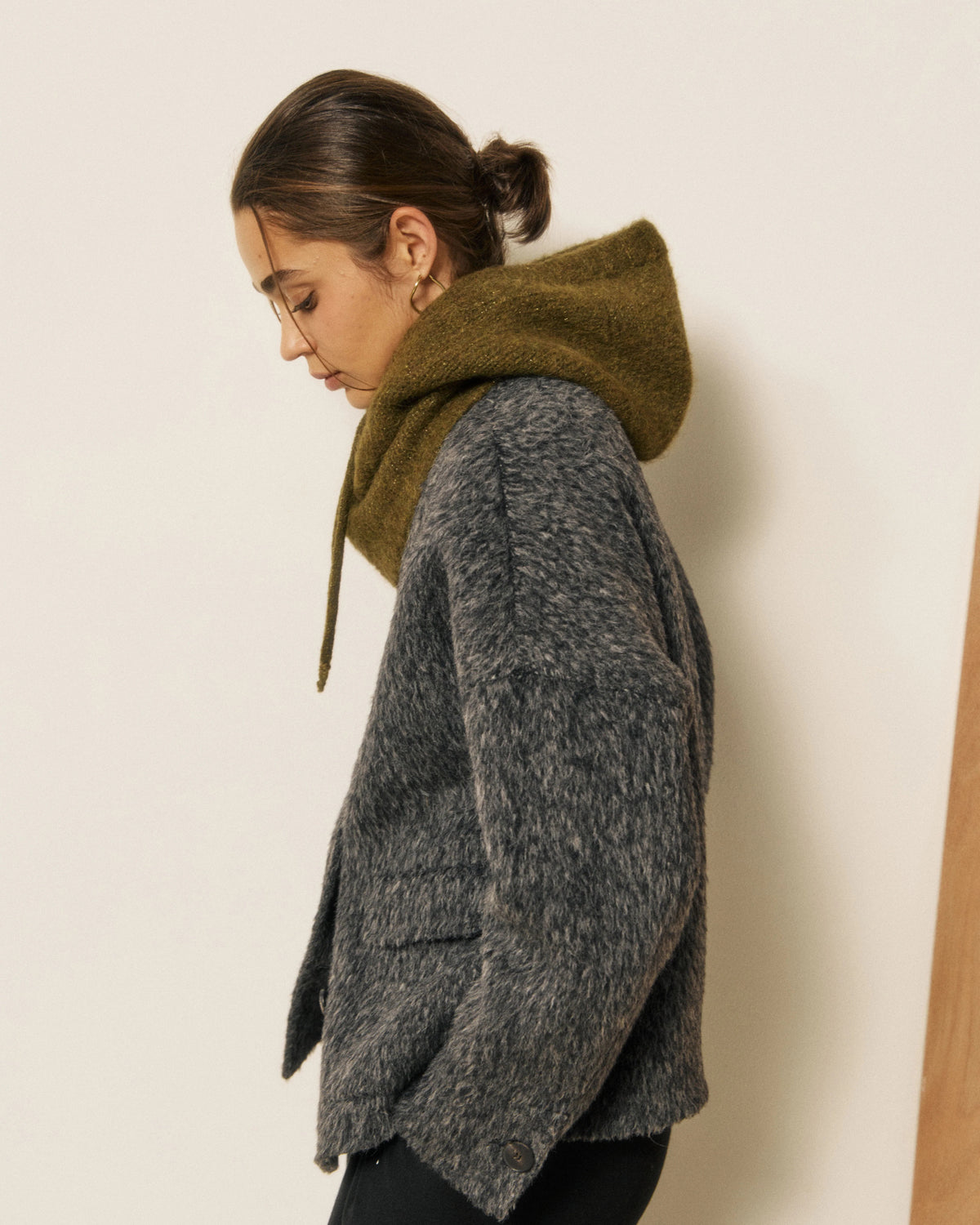 A woman wearing a fuzzy gray coat and the HARTFORD - MEVA HOOD IN KHAKI by HARTFORD—a cozy, merino wool balaclava crafted in Italy with drawstrings—looks directly at the camera against a plain background, adding both warmth and elegance to her winter style.