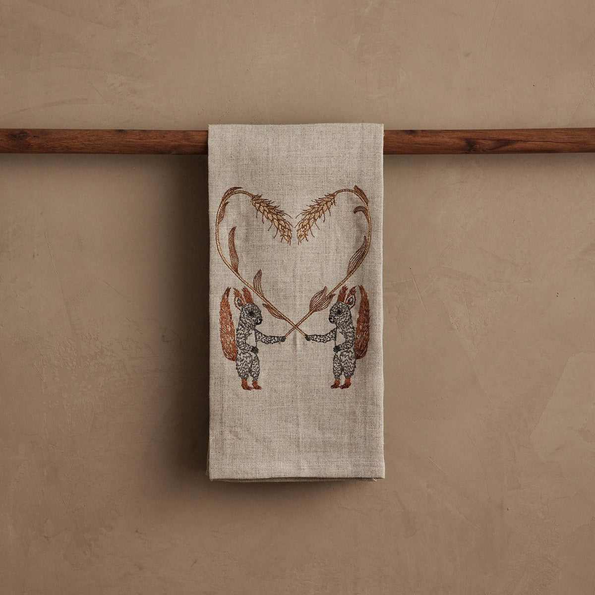 SQUIRRELS WITH WHEAT HEART LINEN TEA TOWEL by CORAL & TUSK featuring two embroidered squirrels holding stalks of wheat that form a heart shape above them.