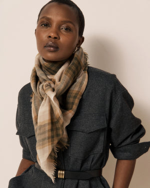 A person wearing a dark belted jumpsuit and the HARTFORD - CHECKED WOOL SCARF IN TAUPE by HARTFORD, crafted in India, stands by a window, gazing confidently at the camera in soft natural light.