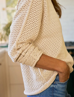 A woman in a sunlit kitchen smiles, wearing the FRANK & EILEEN Monterey Pointelle Rolled Funnel Neck Cotton Sweater in Sand with blue jeans; its machine washable design brings comfort and ease to her day.
