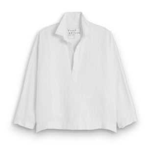 A woman with long brown hair smiles outdoors, wearing the FRANK & EILEEN Gabi Popover Shirt in washed linen white. Green leafy plants and wicker furniture are visible in the background.