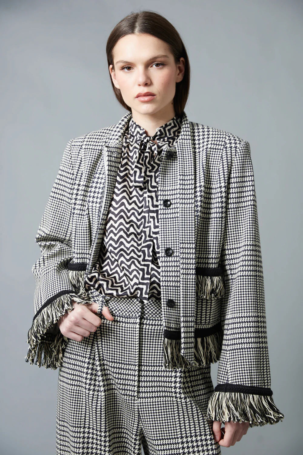 A woman with straight brown hair faces away in a HOPE FOR FLOWERS Tie Neck Blouse—black and white, vertically striped Tencel with puffed sleeves—paired with wide-leg plaid trousers. The plain gray background reflects sustainable style.