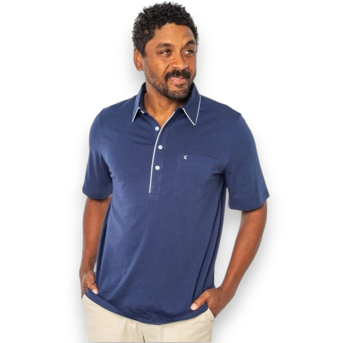 A man with a gray beard is wearing the CRIQUET Throwback Players Shirt in Navy, featuring a classic fit with white trim on the collar and a front pocket, as he stands outdoors surrounded by greenery.
