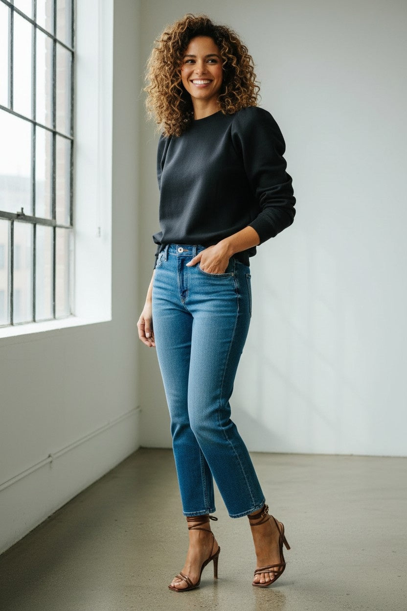 A woman with curly hair, dressed in the KMJ - COOPER SWEATSHIRT IN BLACK by KMJ, blue jeans, and brown heels stands smiling near a large window in a bright, minimalist room.