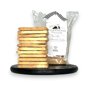 A stack of buttery and crisp vanilla shortbread cookies is displayed on a black serving board in front of a clear package labeled "Fearrington House Signature Collection - Housemade Vanilla Shortbread Cookies" with a black and white striped ribbon tie at the top. These delectable treats could easily be found at The Fearrington House Restaurant.
