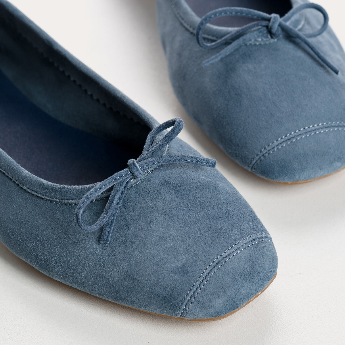 The REQINS Harmony Suede Ballerina Flats in Blue Jean feature luxurious goat suede, a delicate front bow, and a low brown heel, displayed on a plain white background.