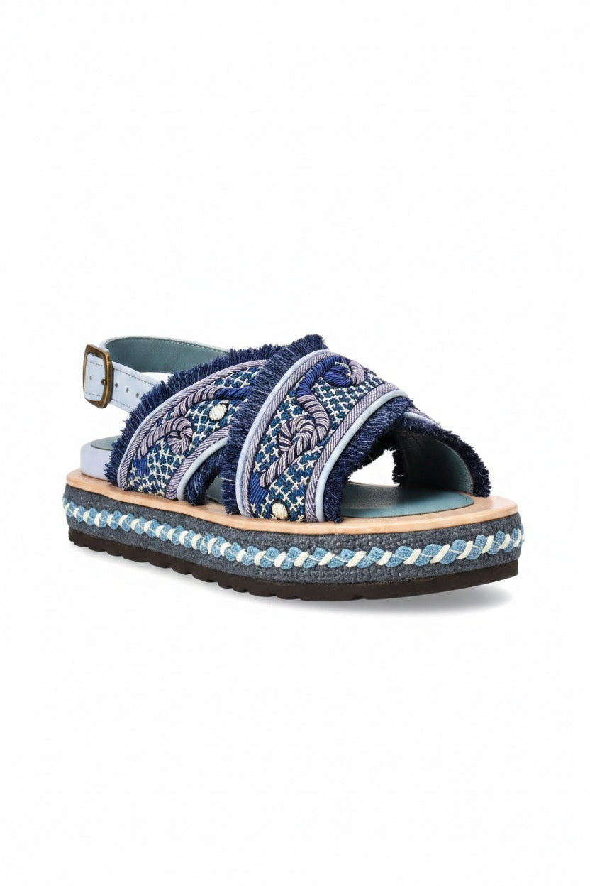 Introducing the MEHER KAKALIA DA GAMMA BLOC SANDAL in Yacht Blue Deep Blue: a chic sandal with blue cross-straps, intricate hand embroidery, frayed edges, and a decorative platform sole featuring blue and white braid accents. It includes an adjustable ankle strap with a gold buckle.