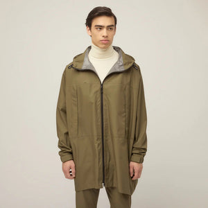 A person models the TILLEY - PACKABLE HOODED PONCHO in olive green, layered over a white turtleneck, standing against a plain light background and gazing to the side with a neutral expression. Brand: TILLEY.