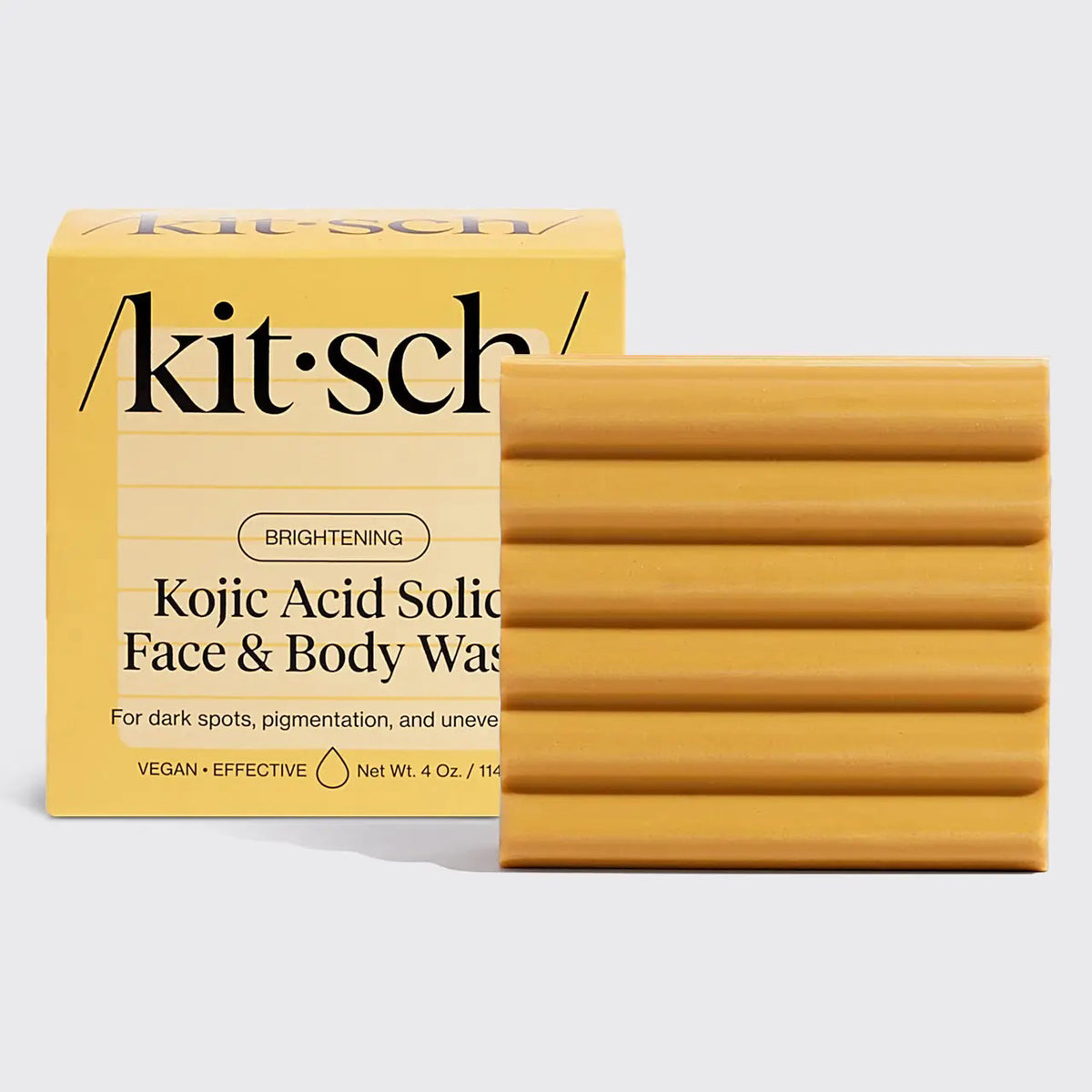 A ribbed, rectangular bar of soap is next to a yellow box labeled "KITSCH - KOJIC ACID FACE AND BODY BAR." It includes turmeric to address dark spots, pigmentation, and uneven skin tone. Net weight: 4 oz.