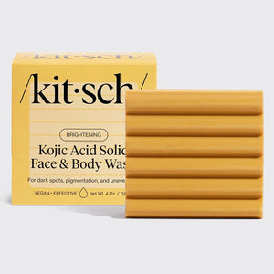 A ribbed, rectangular bar of soap is next to a yellow box labeled "KITSCH - KOJIC ACID FACE AND BODY BAR." It includes turmeric to address dark spots, pigmentation, and uneven skin tone. Net weight: 4 oz.