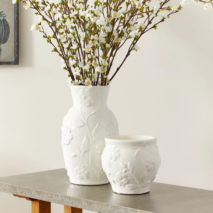 The NAPA HOME AND GARDEN - BLOSSOM CACHEPOT is a white ceramic vase ideal for home decor. It boasts a stunning floral design with embossed leaves and flowers, featuring a rounded shape and slightly flared top edge, reminiscent of a graceful blossom.