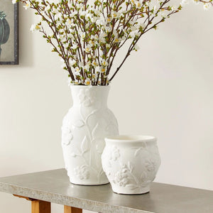 The NAPA HOME AND GARDEN - BLOSSOM CACHEPOT is a white ceramic vase ideal for home decor. It boasts a stunning floral design with embossed leaves and flowers, featuring a rounded shape and slightly flared top edge, reminiscent of a graceful blossom.