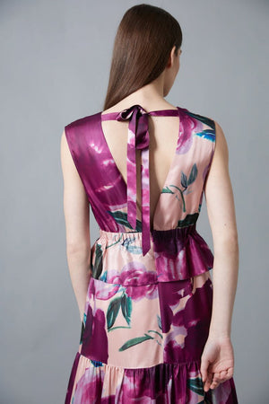A woman with straight brown hair and a neutral expression wears the HOPE FOR FLOWERS Tiered Maxi Dress, a sleeveless, floor-length style featuring bold purple and pink floral print, standing against a gray background.
