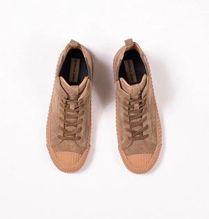 A pair of DANIELLA LEHAVI - OSAKA HIGH TOP SNEAKERS with a contemporary design, featuring brown suede uppers and rugged rubber soles, are placed side by side on a white pedestal against a plain beige background. These comfortable high-top sneakers boast a lace-up design and ribbed toe caps for added durability.