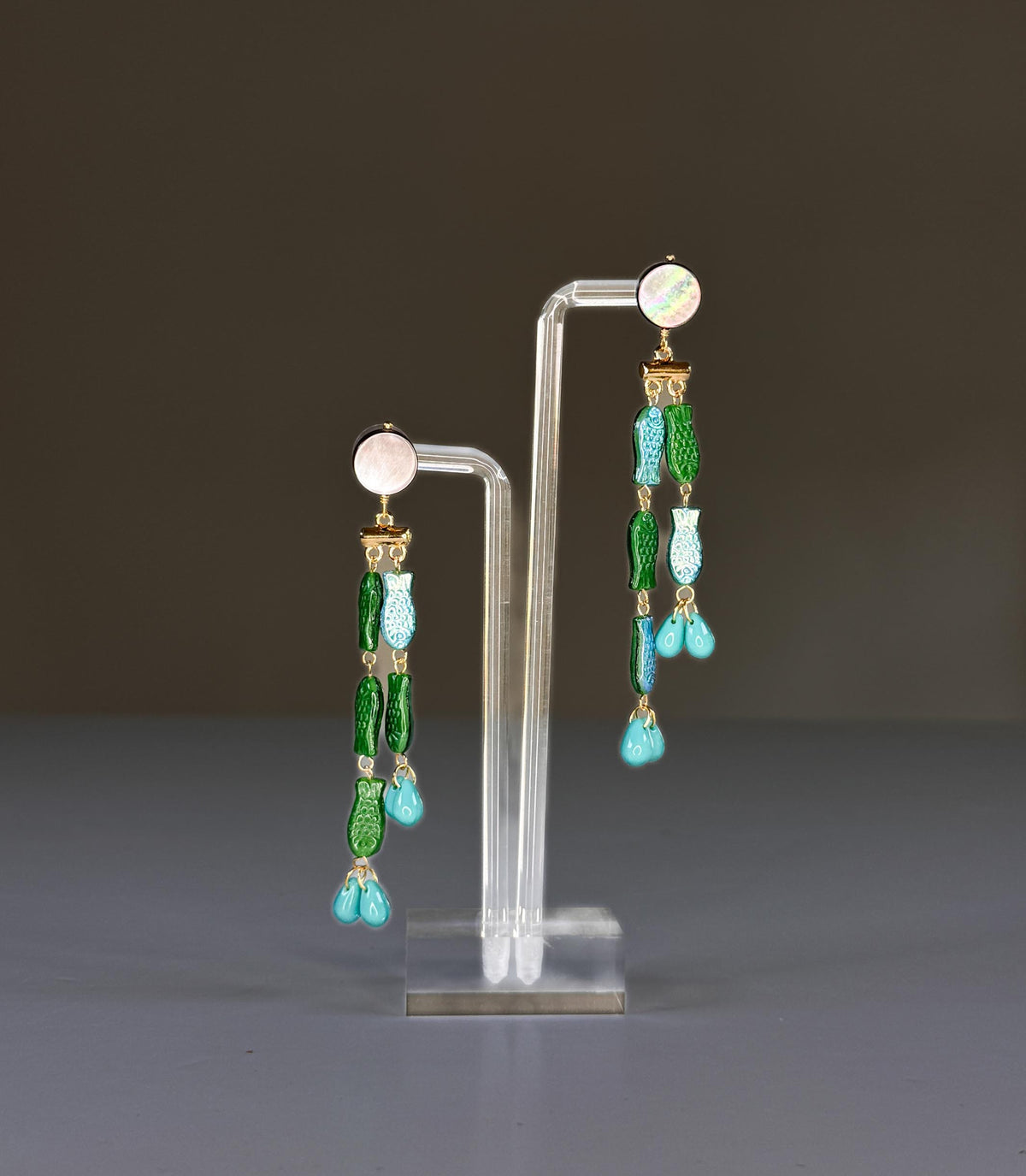 The SANDRINE FRANCE STUDIO - EVIE EARRINGS feature handmade round mother-of-pearl studs, gold connectors, green and blue fish charms, and turquoise teardrop beads at the ends.