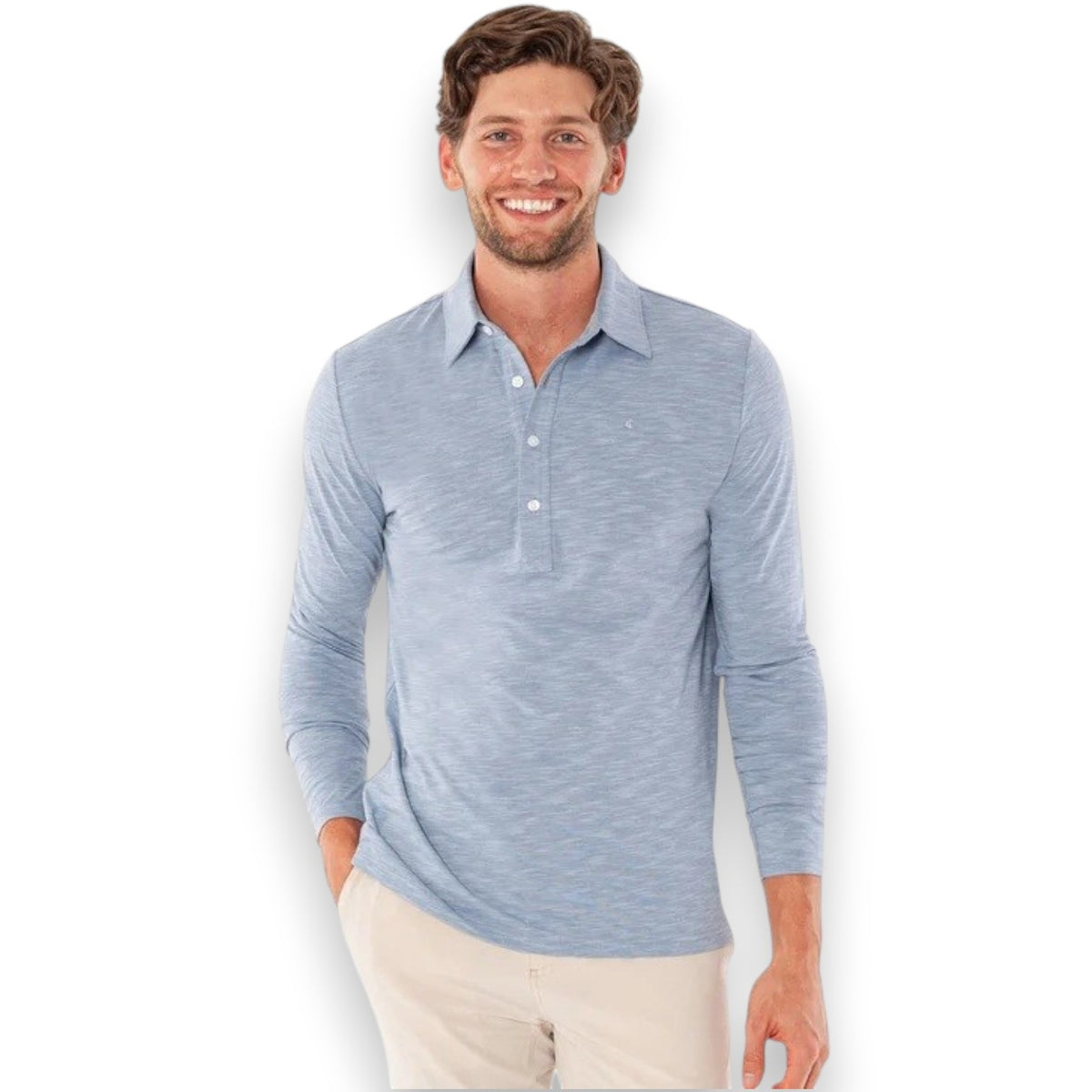 A man wearing the CRIQUET BRRR° Long Sleeve Range Polo in Bluestone and a green cap smiles while holding a can, leaning against a mossy tree with a cabin and rocks in the background.