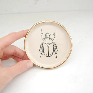 The COMPOST DESIGN - BEETLE DISH 4" by COMPOST DESIGN showcases a black beetle illustration at its center, surrounded by a 22k gold rim. Set on white, it features an abstract shell design background—perfect for decoration or as part of a treasured collection.