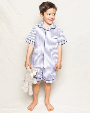 A young boy stands barefoot against a plain background, smiling and looking off to the side. He is wearing an elegant PETITE PLUME FEARRINGTON LIFESTYLE COLLECTION CHILDREN'S TWILL NAVY FRENCH TICKING PAJAMA SHORT SET and is holding a plush grey teddy bear by its arm.