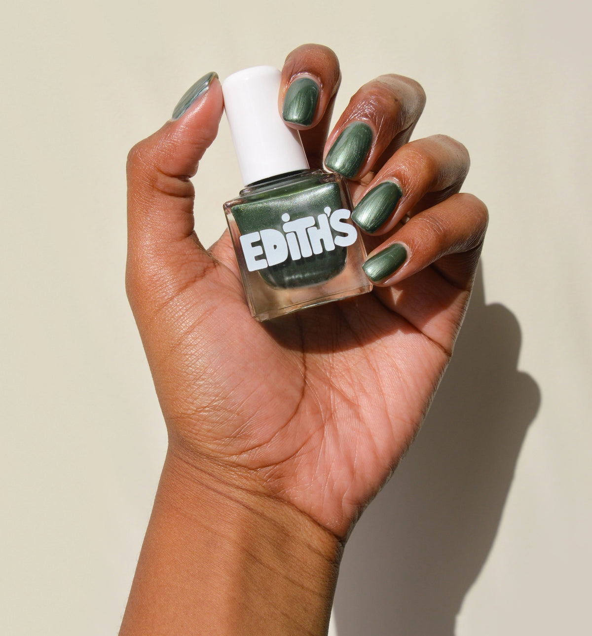 A hand with dark skin holds a bottle of TENOVERTEN - KIND OF A BIG DILL NAIL POLISH by TEN OVER TEN. The nails are painted in the same metallic green shade, set against a light, neutral background.